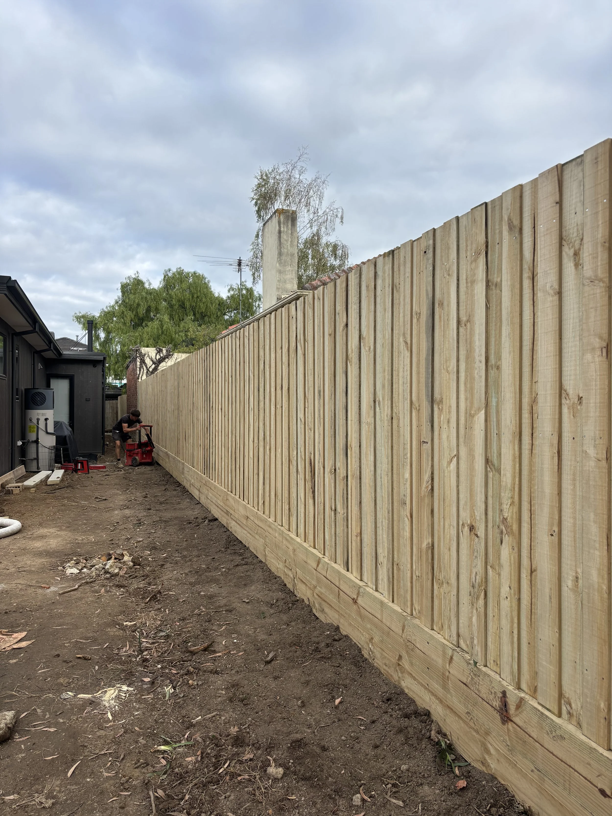 A backyard with a newly built wooden fence, dirt ground, and a person working on construction near a black building.