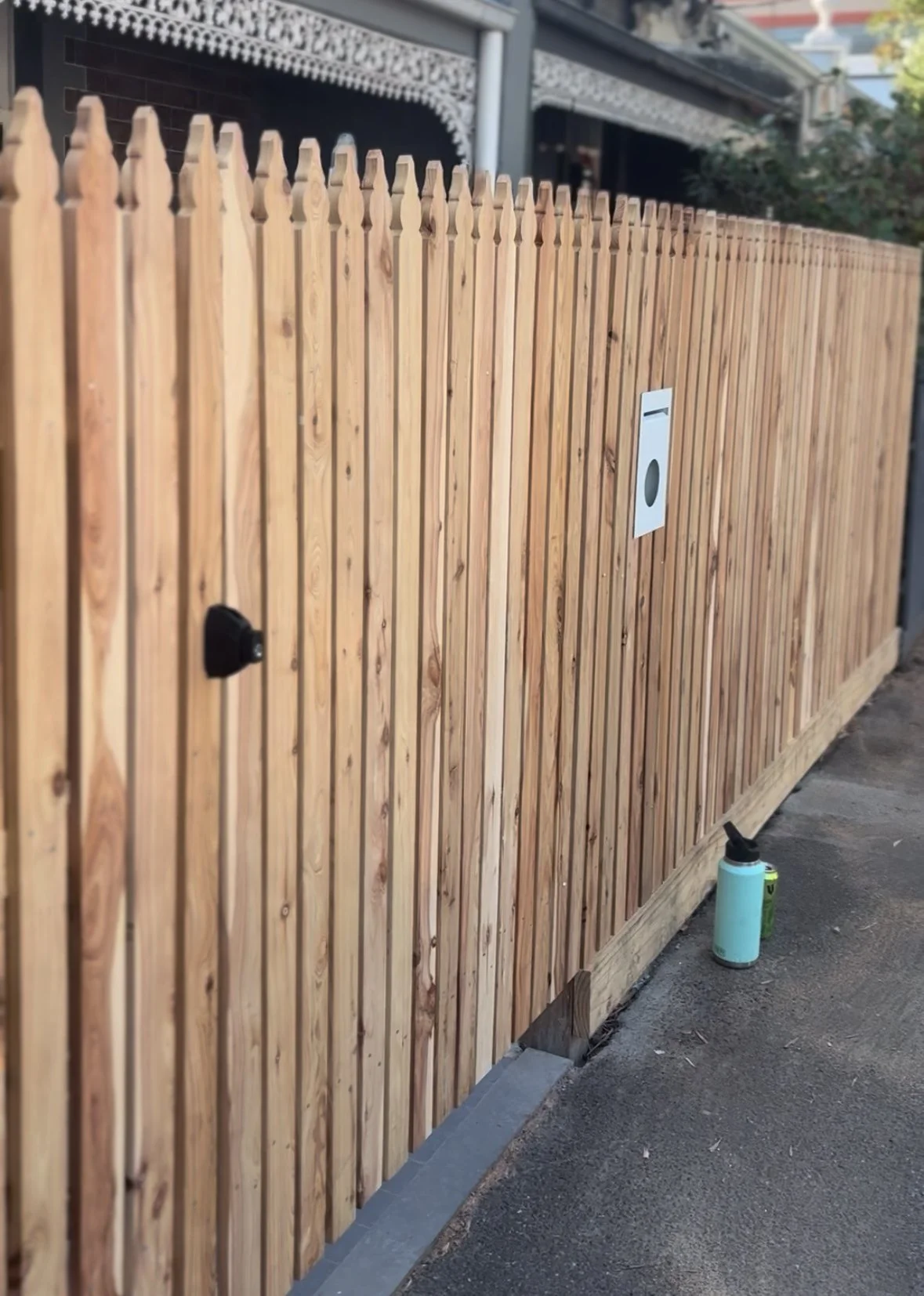 Wooden privacy fence with mail slot and lock, on a paved sidewalk with a water bottle and spray can on the ground nearby.