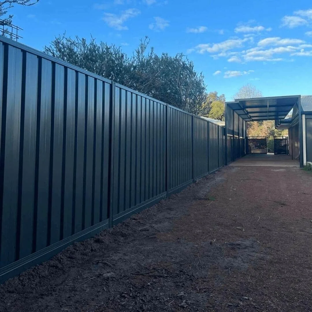 A black metal fence running alongside a dirt pathway with a covered area and gate in the distance, under a partly cloudy blue sky.