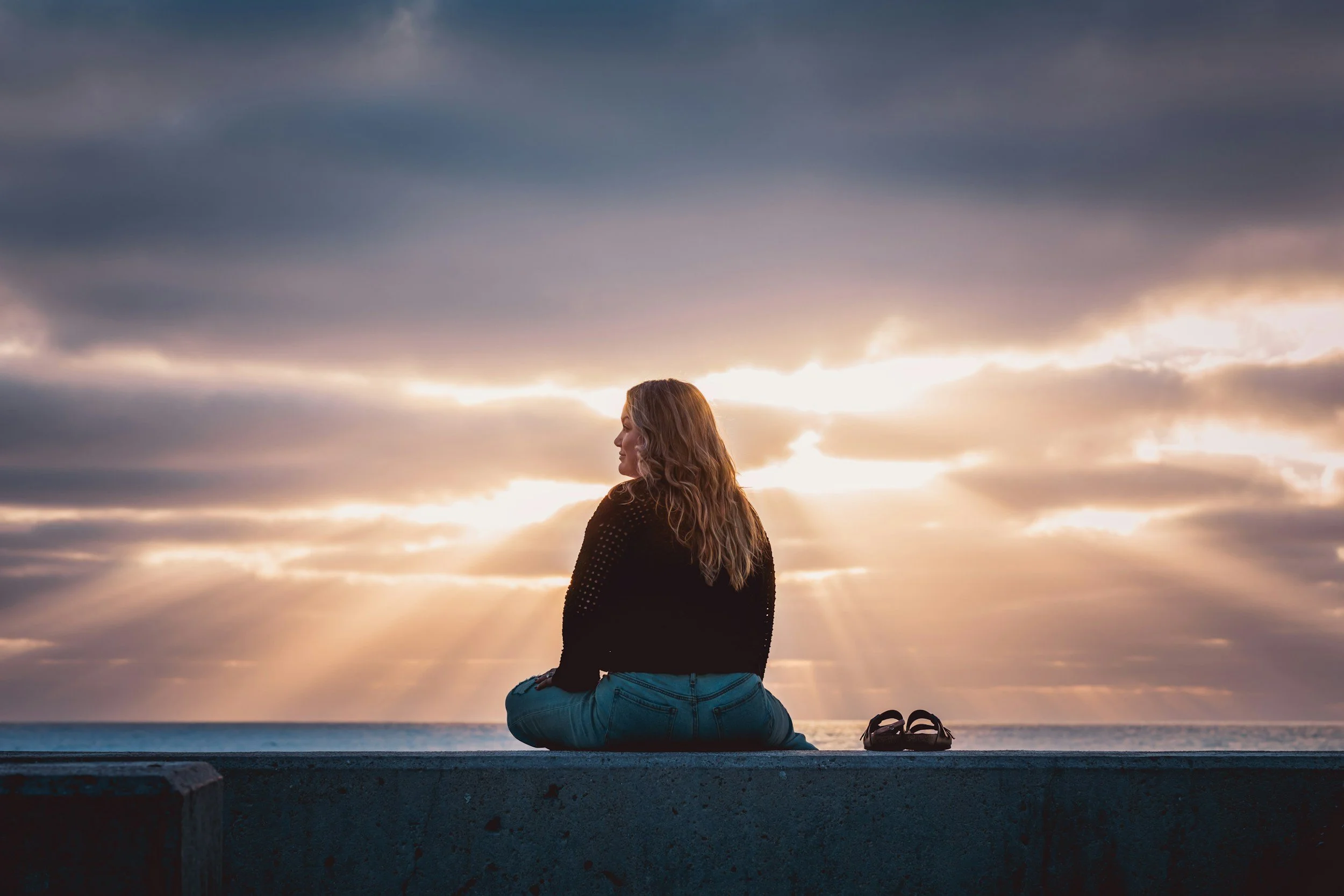 women with her back turned, sitting on a wall by the beach watching the sunset