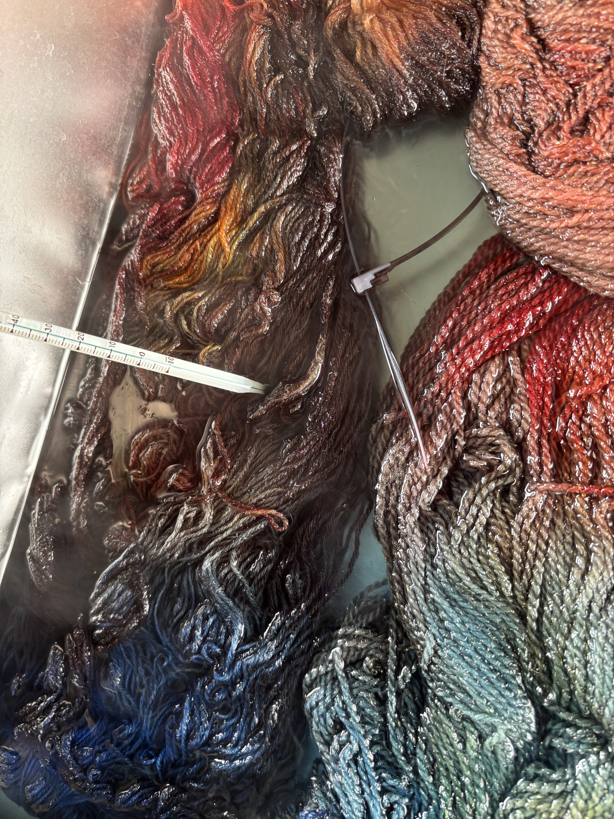 Close-up image of multicolored silk threads, ranging from red at the top, blending into orange, brown, blue, and green, with a needle and a small container of liquid dye in the background for dyeing the silk.