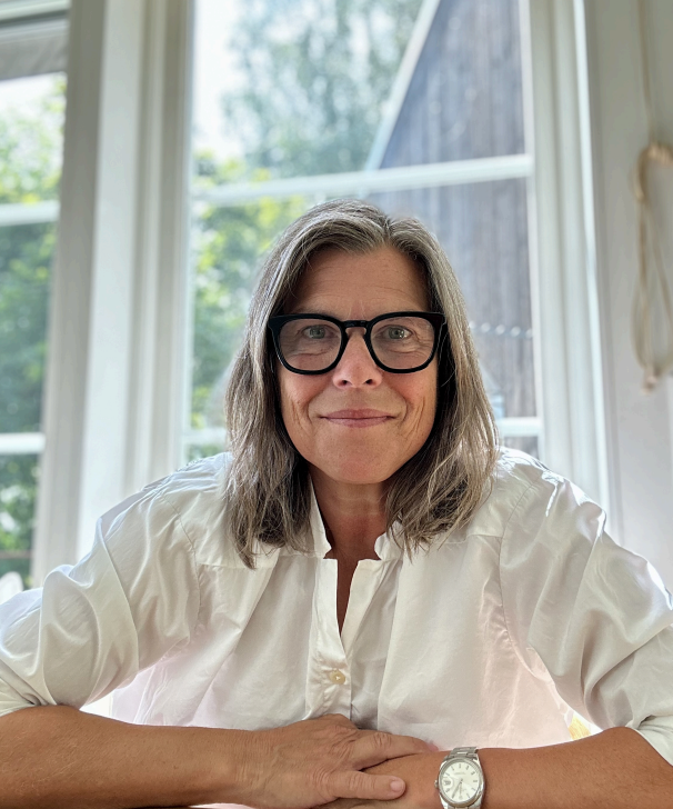 A woman with shoulder-length gray hair, wearing glasses and a white shirt, sitting at a table with a window and green trees in the background.