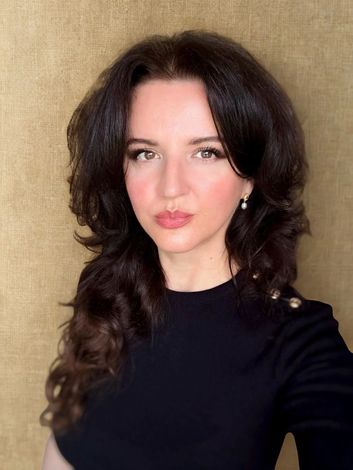 Porträt einer jungen Frau mit schwarzen, lockigen Haaren und goldenen Ohrringen, vor einer beige hintergrund. Sie trägt ein schwarzes Oberteil.