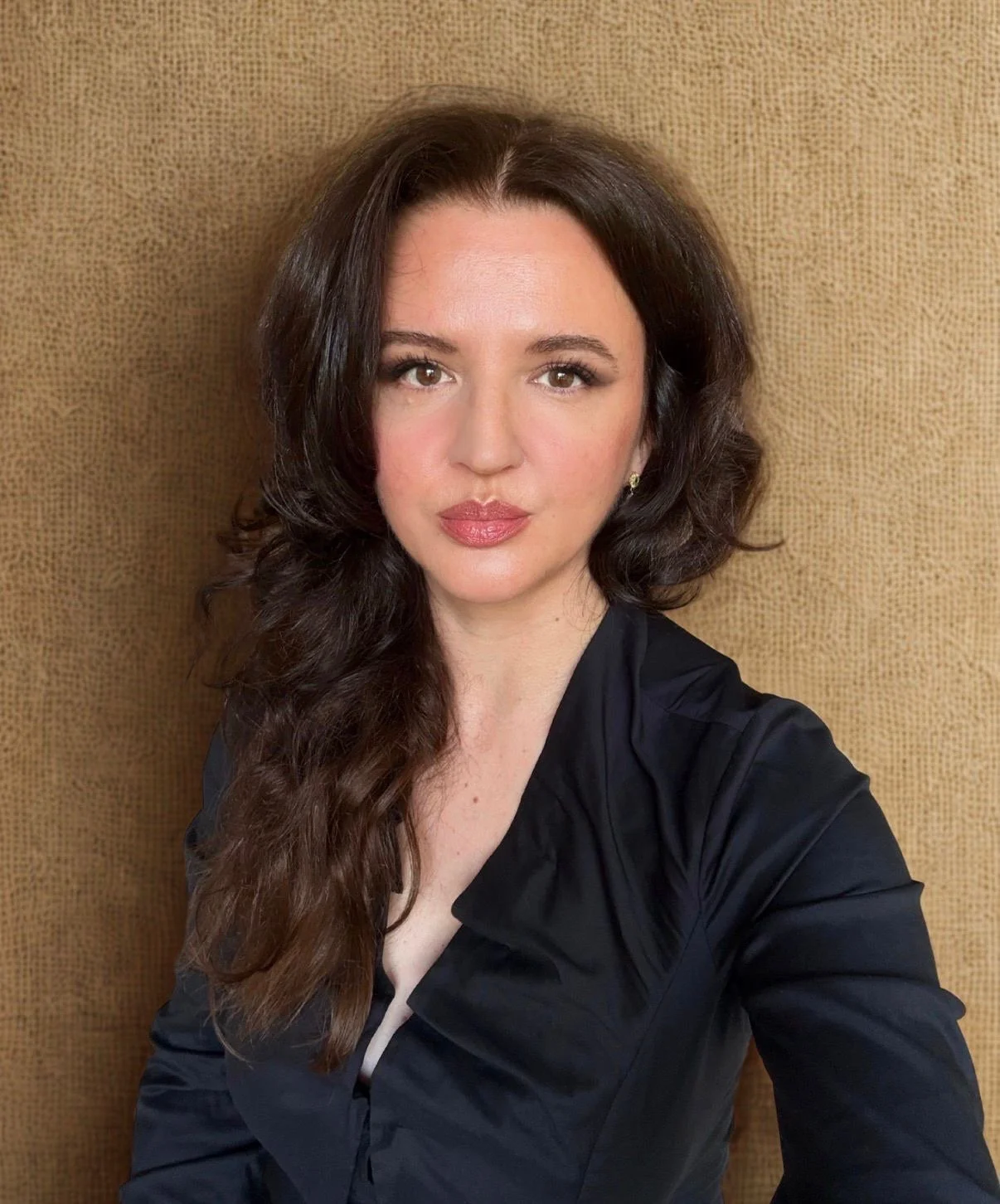 Porträt einer jungen Frau mit braunen, gewellten Haaren und rotem Lippenstift vor einem braunen Hintergrund.