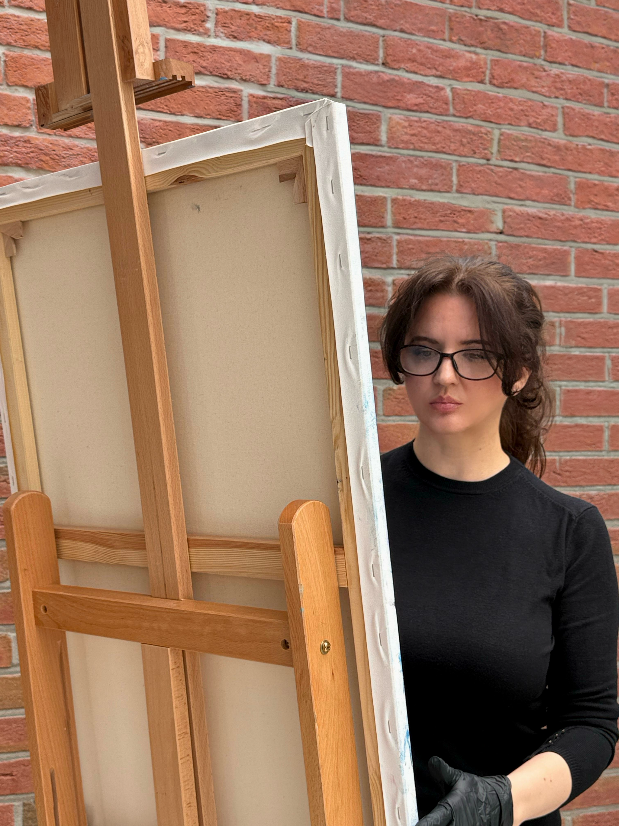 Eine junge Frau mit dunklen, lockigen Haaren, Brille und schwarzer Kleidung steht vor einem Holzrahmen und einer Leinwand, die auf einer Unterlage liegt, vor einer roten Backsteinwand.