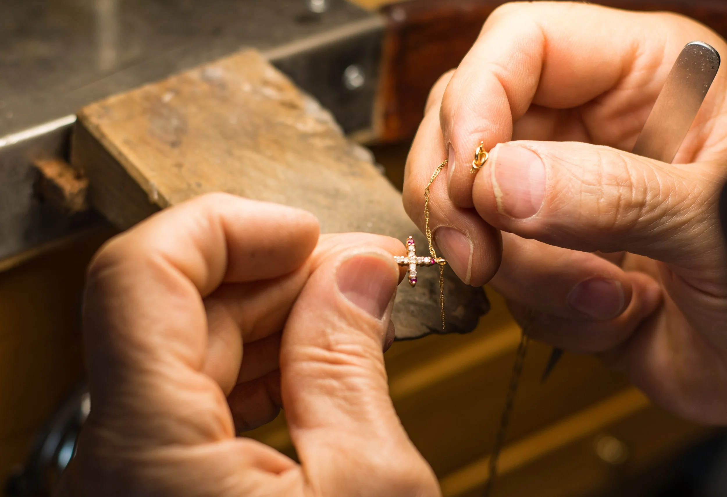 A person working on a gold necklace with a cross pendant, holding the chain with one hand and adjusting the pendant with the other, in a jewelry workshop.