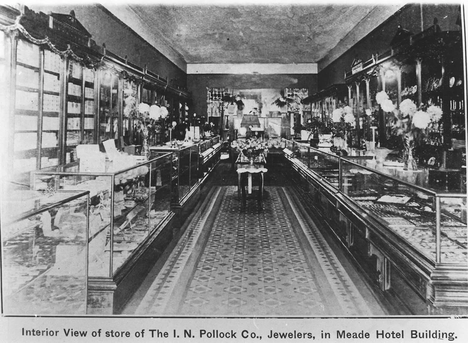 Black and white interior view of a jewelry store named The I. N. Pollock Co., located in the Meade Hotel Building. The store features glass display cases along both sides filled with jewelry and decorative items, with floral arrangements placed on top. A patterned floor leads to a distant back wall.