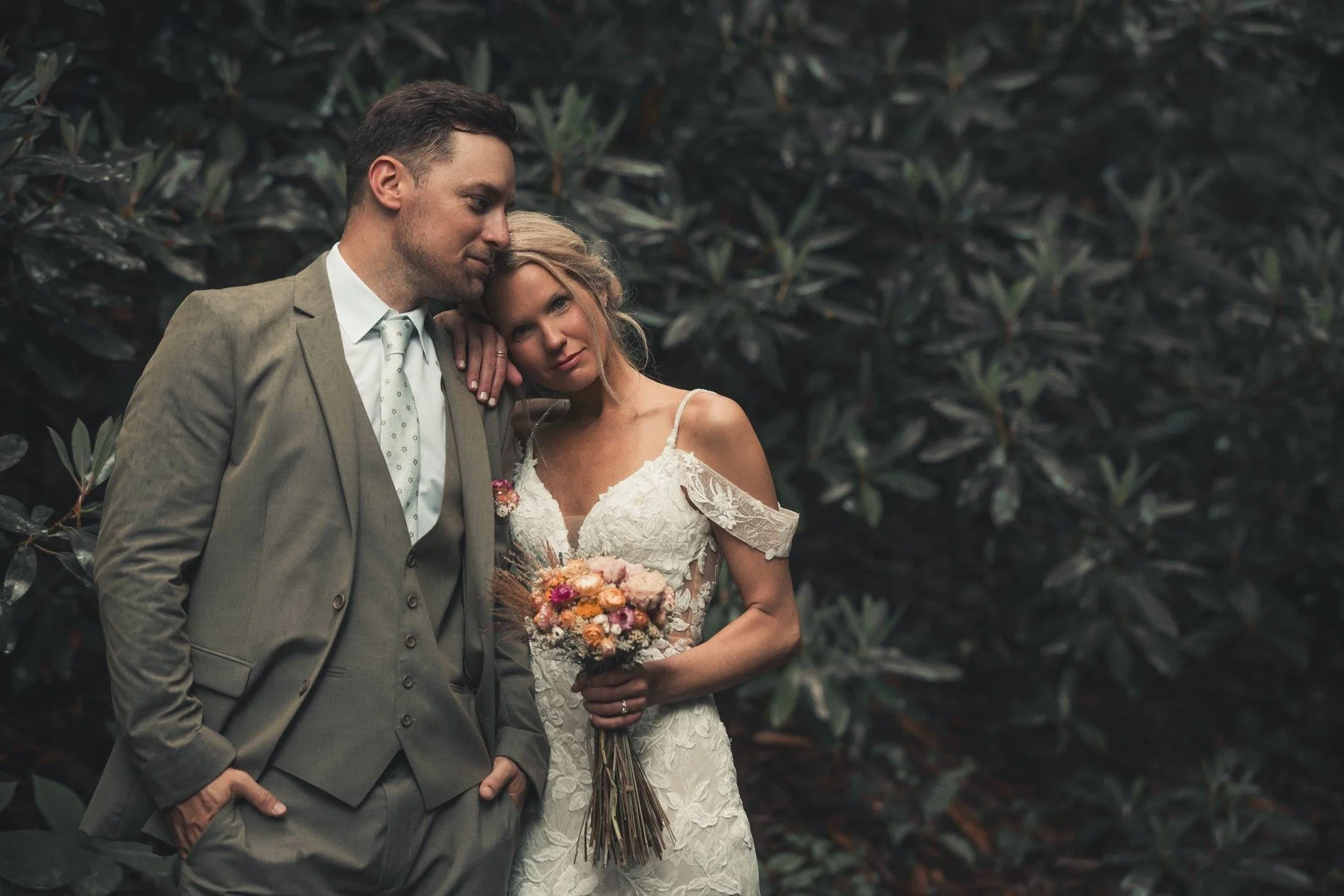 A bride and groom standing close together outdoors, with the bride holding a bouquet of flowers and resting her head on the groom's shoulder. The groom is wearing a light gray suit with a white shirt and patterned tie, and the bride is in a lace off-shoulder wedding dress with light-colored hair styled in loose waves. They are surrounded by dark green foliage.