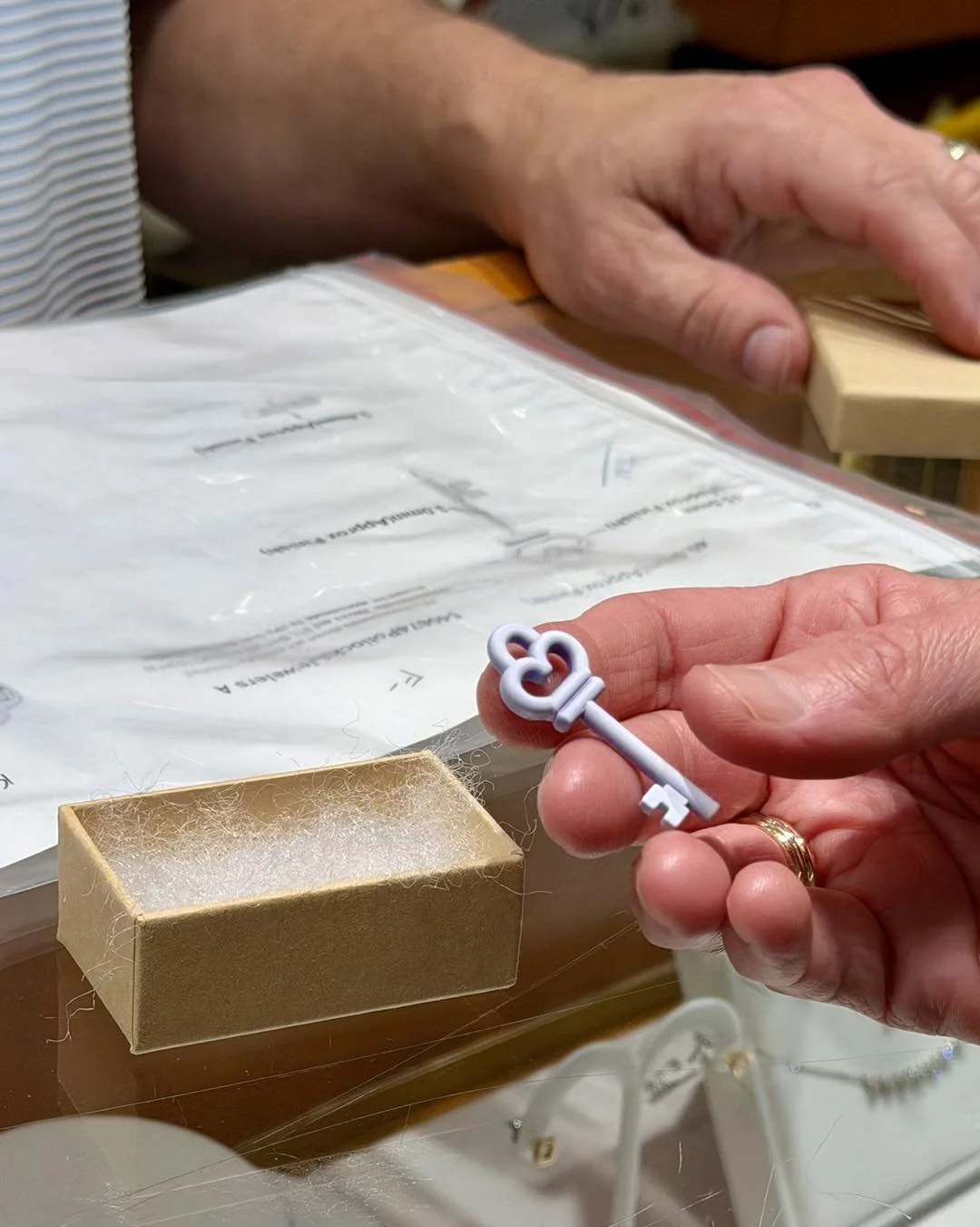 A person holding a small white decorative key with a heart-shaped bow, near a tiny cardboard box on a glass surface. In the background, another person’s hand is visible near a plastic bag with papers or packaging.