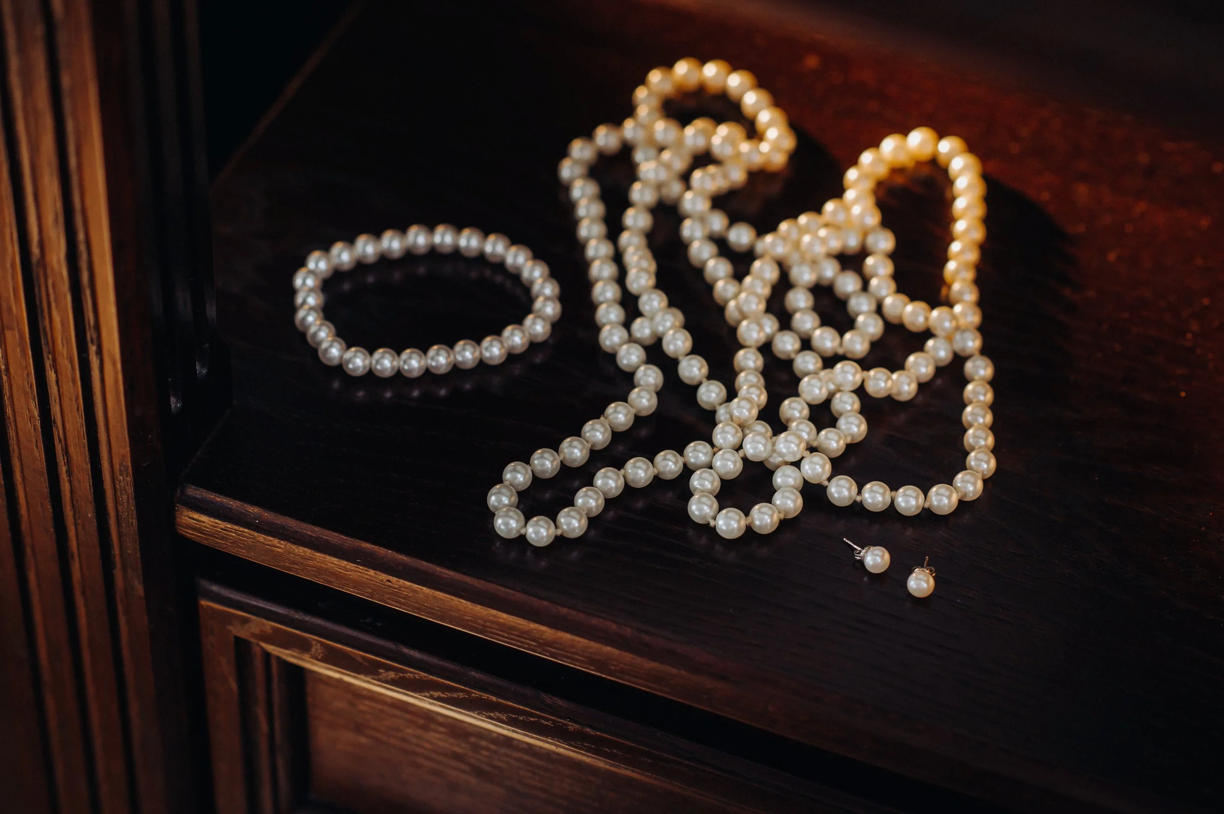 A string of white pearl necklaces and a bracelet, along with matching pearl earrings, are displayed on a dark wooden surface.