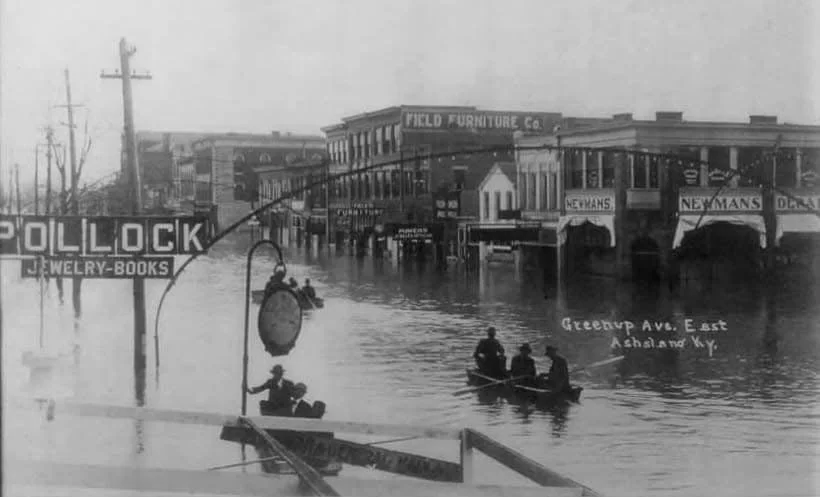 Historical black and white photo of a flooded street in Ashland, Kentucky, showing buildings, storefronts, and people rowing boats on the water.