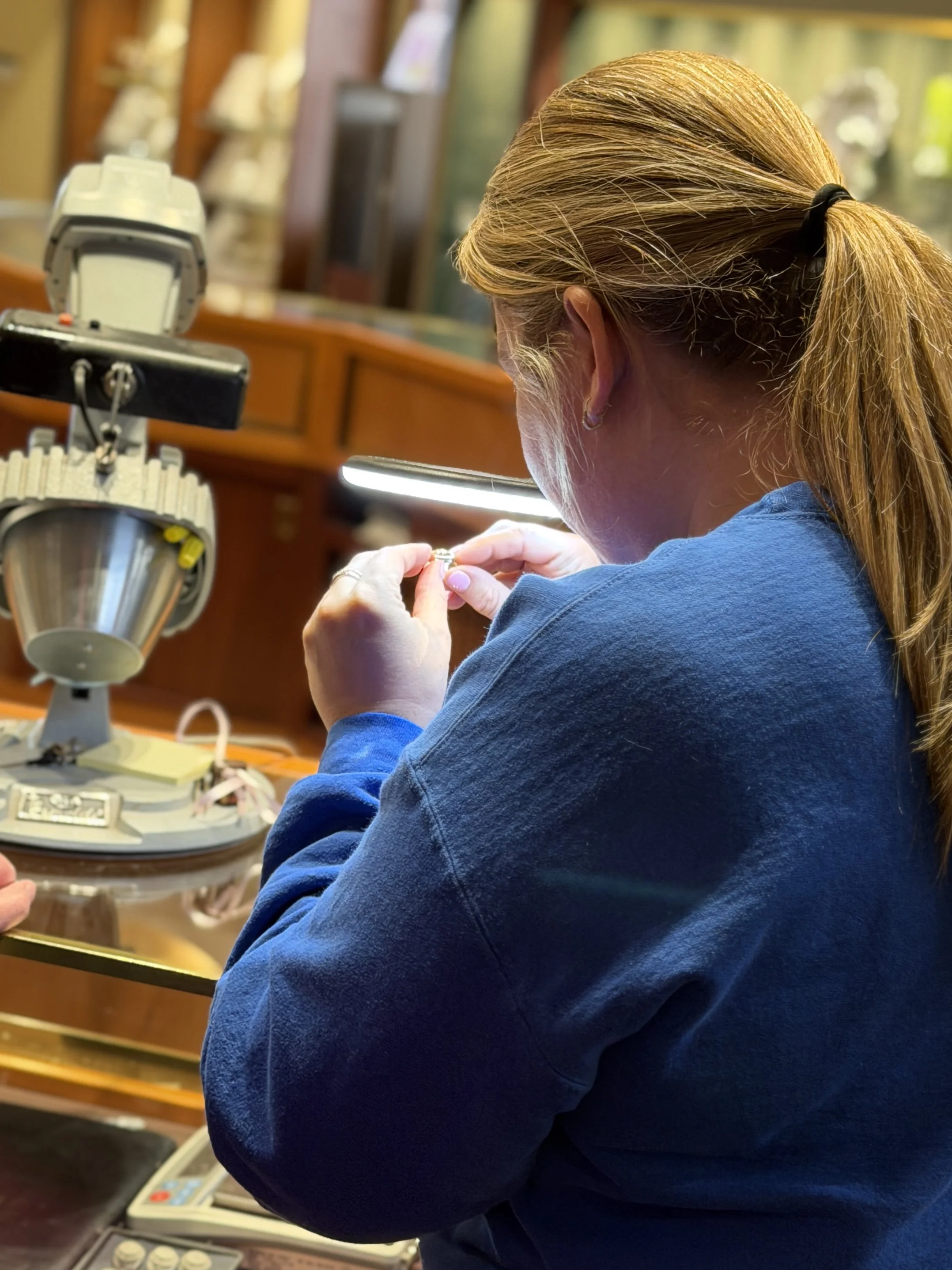A woman with red hair tied back in a ponytail working with jewelry at a jewelry bench. Various tools and equipment are visible on the bench.