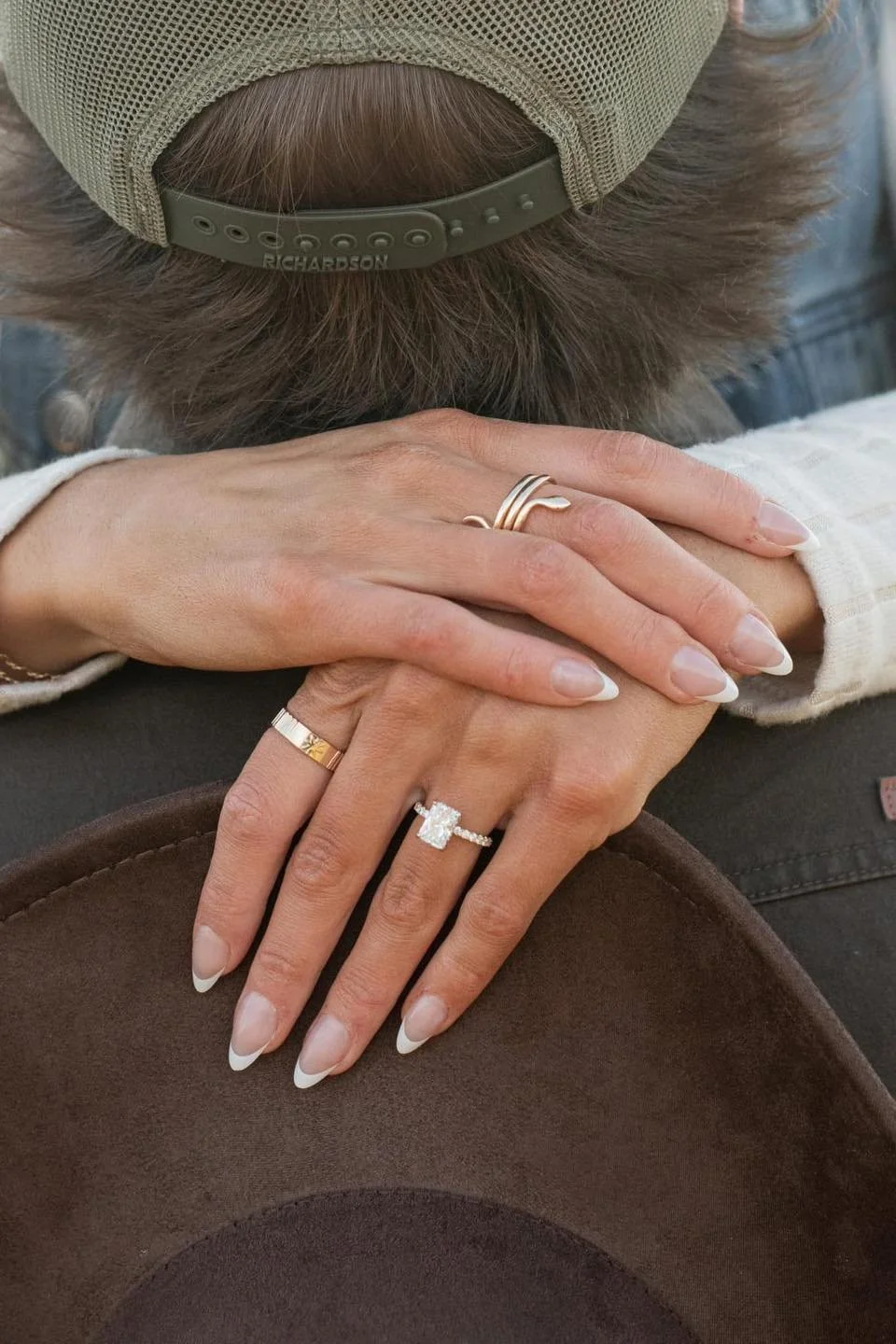 Close-up of two hands with rings resting on a brown hat surface. The hand on top has a gold band, while the hand underneath has an engagement ring with a large center stone and smaller surrounding stones. Both hands have manicured nails with French tips.