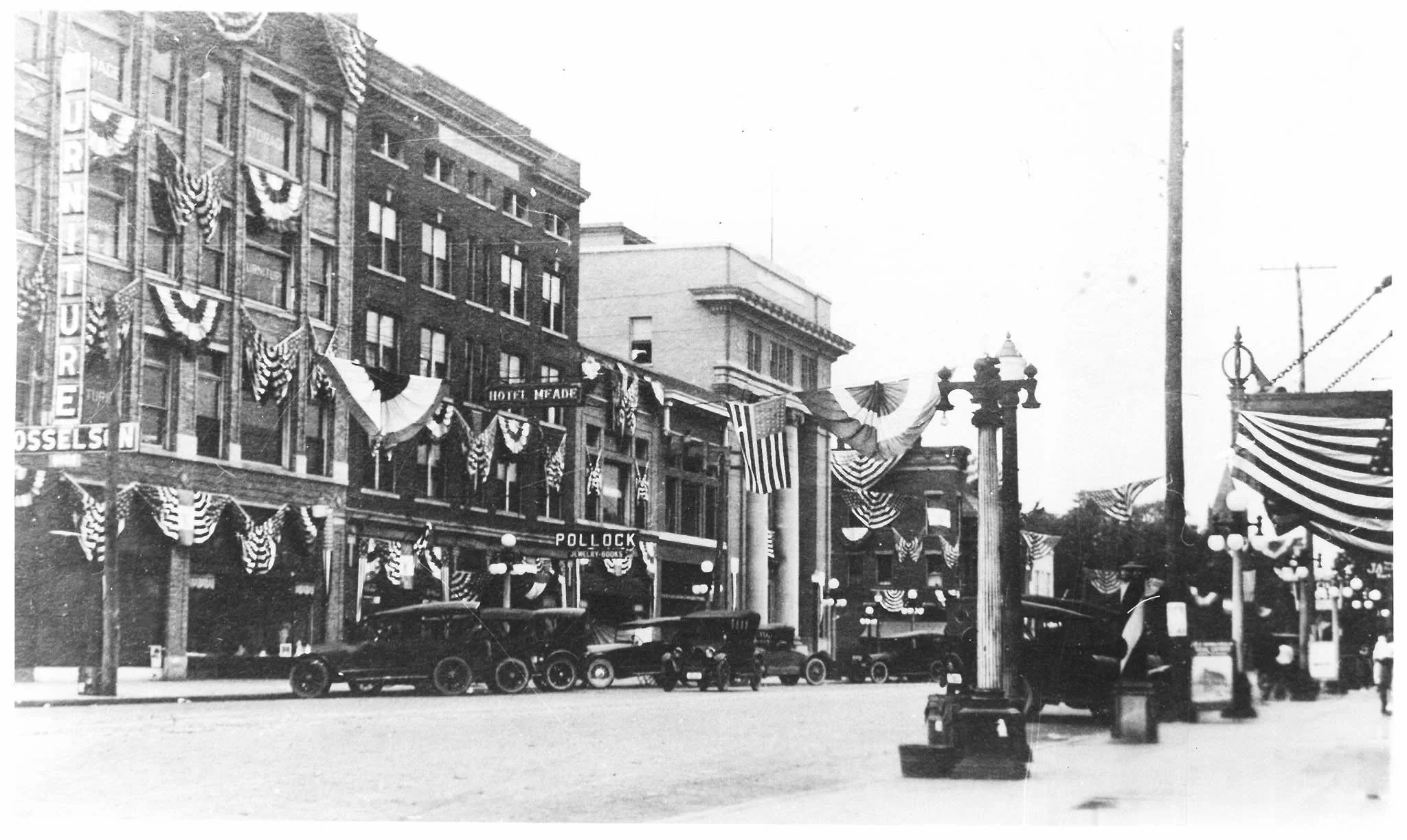 Historical black-and-white street scene decorated with patriotic banners and flags, with parked vintage cars and buildings in the background, celebrating a national holiday.