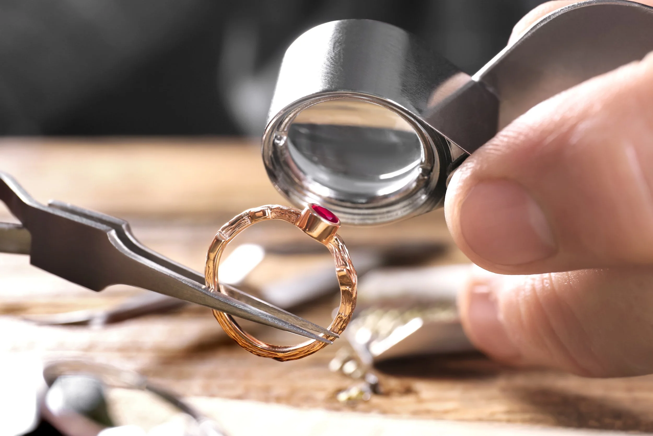 Jeweler using a magnifying glass to examine a gold ring with a red gemstone in a workshop.