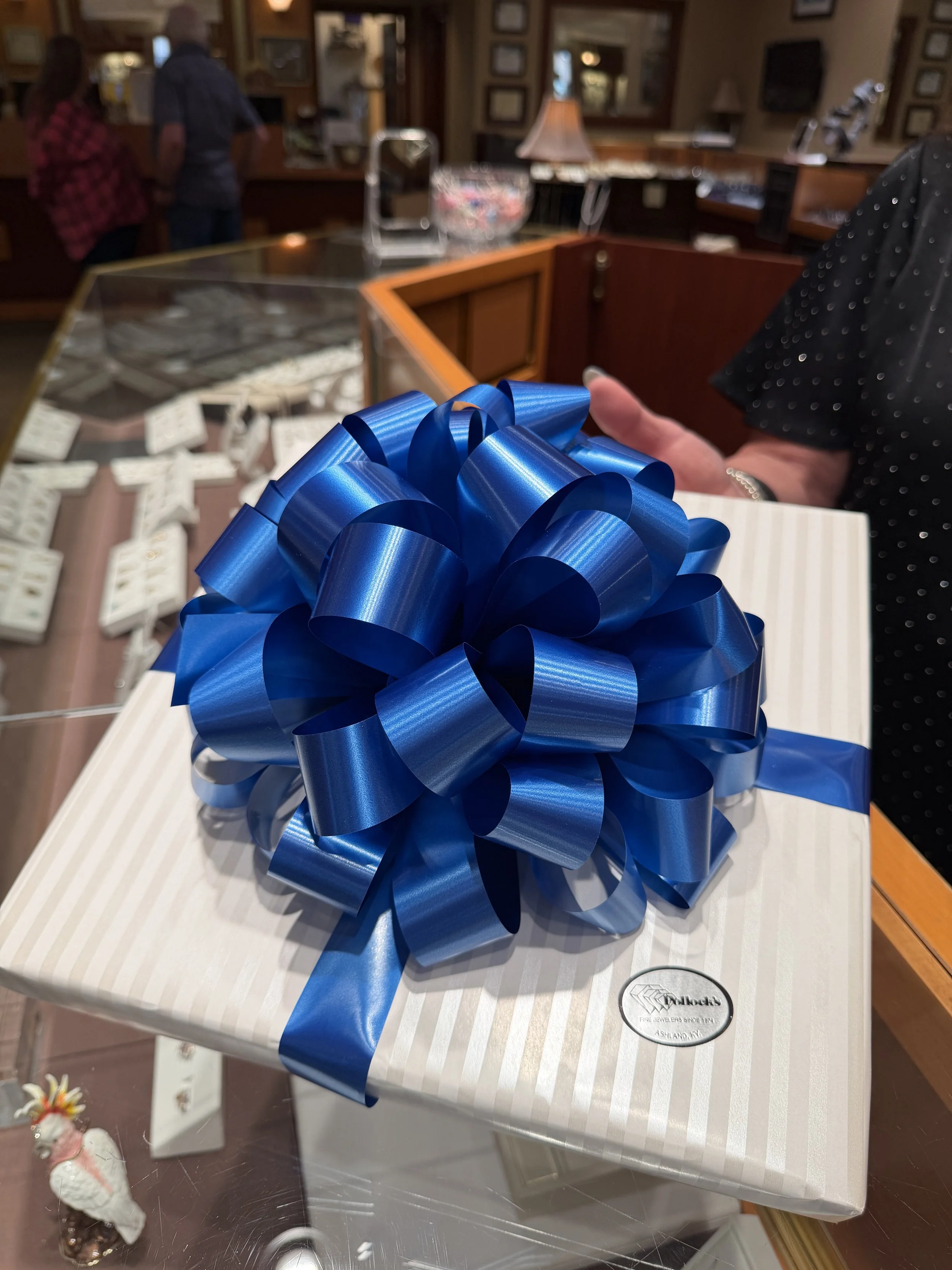 A wrapped gift box with a large, shiny blue ribbon and bow on top, placed on a white table.