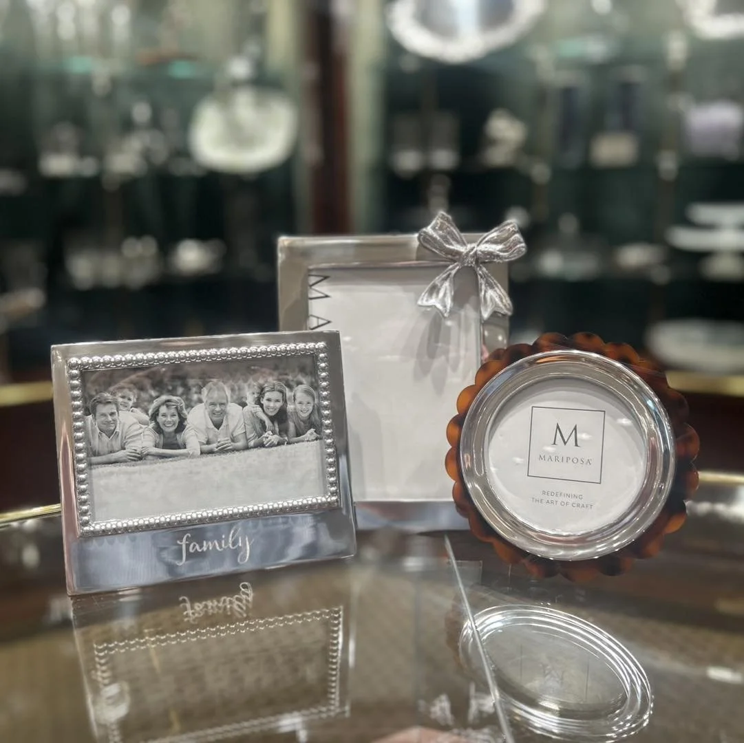 A table display with a black-and-white family photo graphic in a decorative frame, a container with a bow, and a round item with a label that reads 'Mariposa' and 'Redefining the art of craft' in a shop or showroom setting.