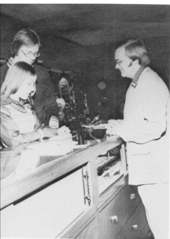 A black and white photo of a man in a white coat talking to three children at a counter, possibly in a bakery or cafe.