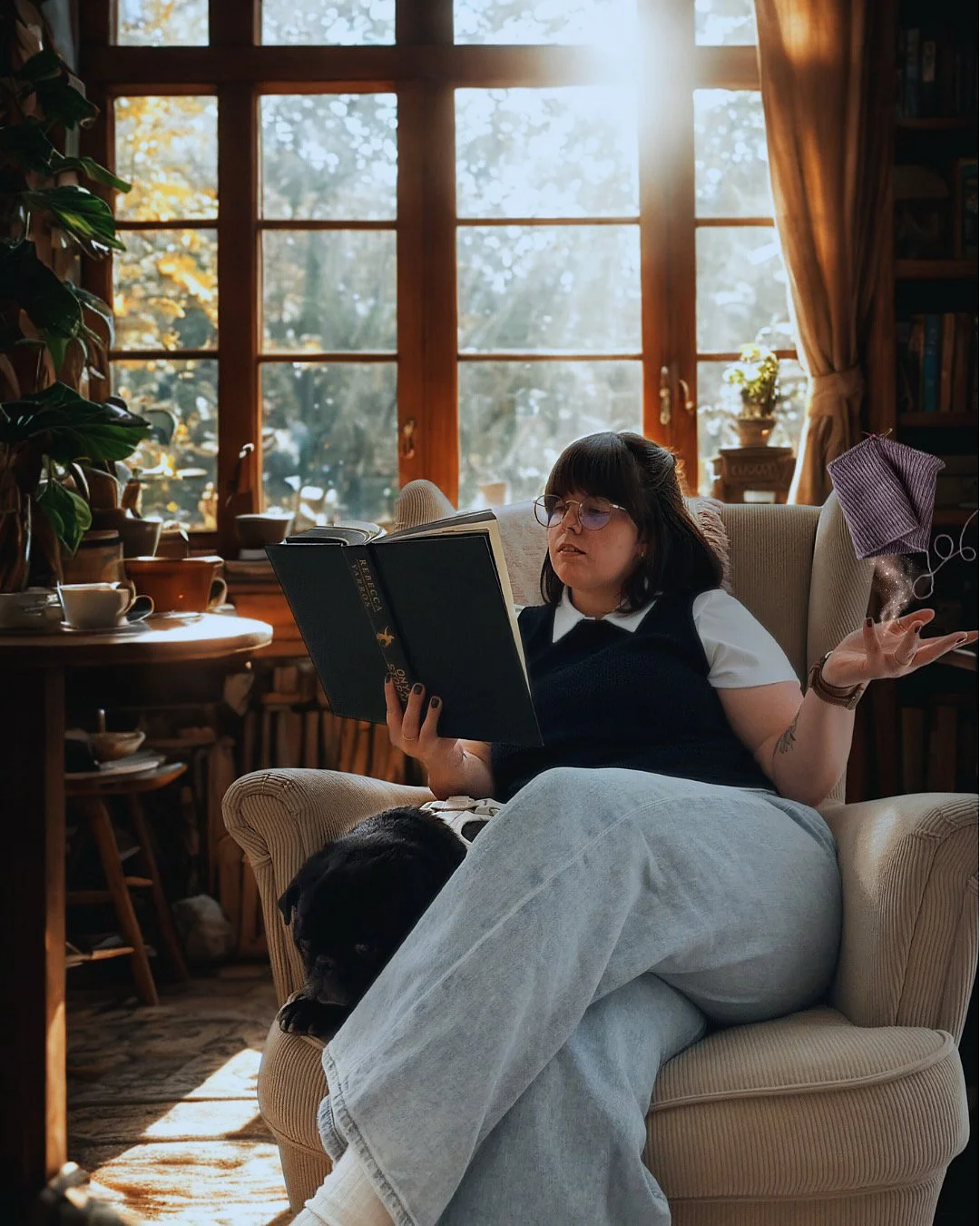 A woman with glasses and dark hair reading a book while sitting on a beige armchair in a cozy sunlit room, with a black dog resting on her lap.