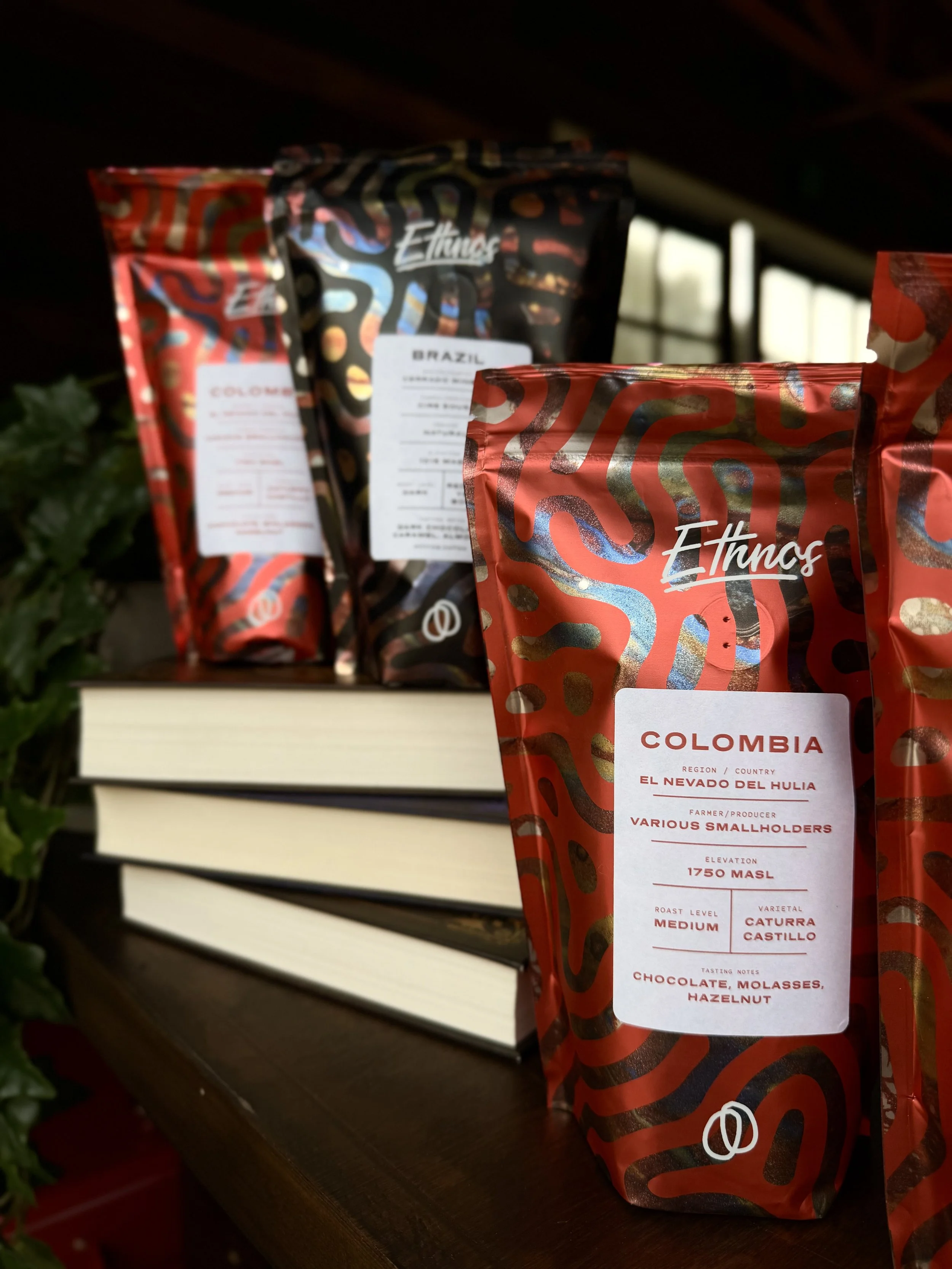 A close-up of coffee bags on a wooden shelf. The bags are decorated with colorful, abstract patterns and have white labels with red text indicating Colombia coffee origin, elevation, roast level, and tasting notes.