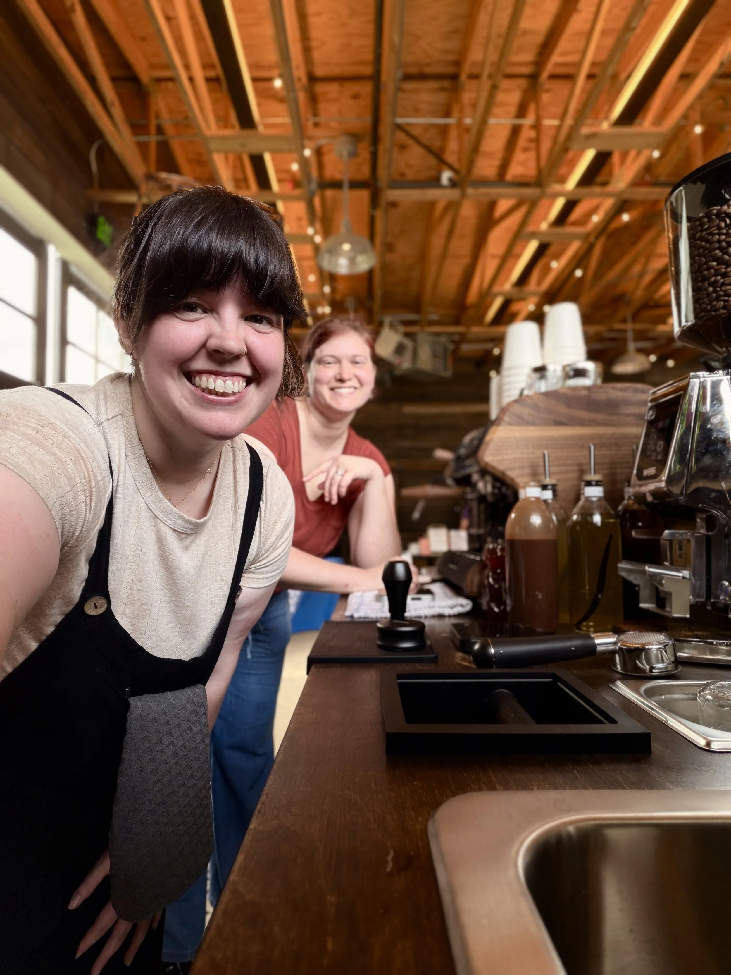 Two smiling women standing behind a coffee counter in a cafe with wooden ceiling and warm lighting.