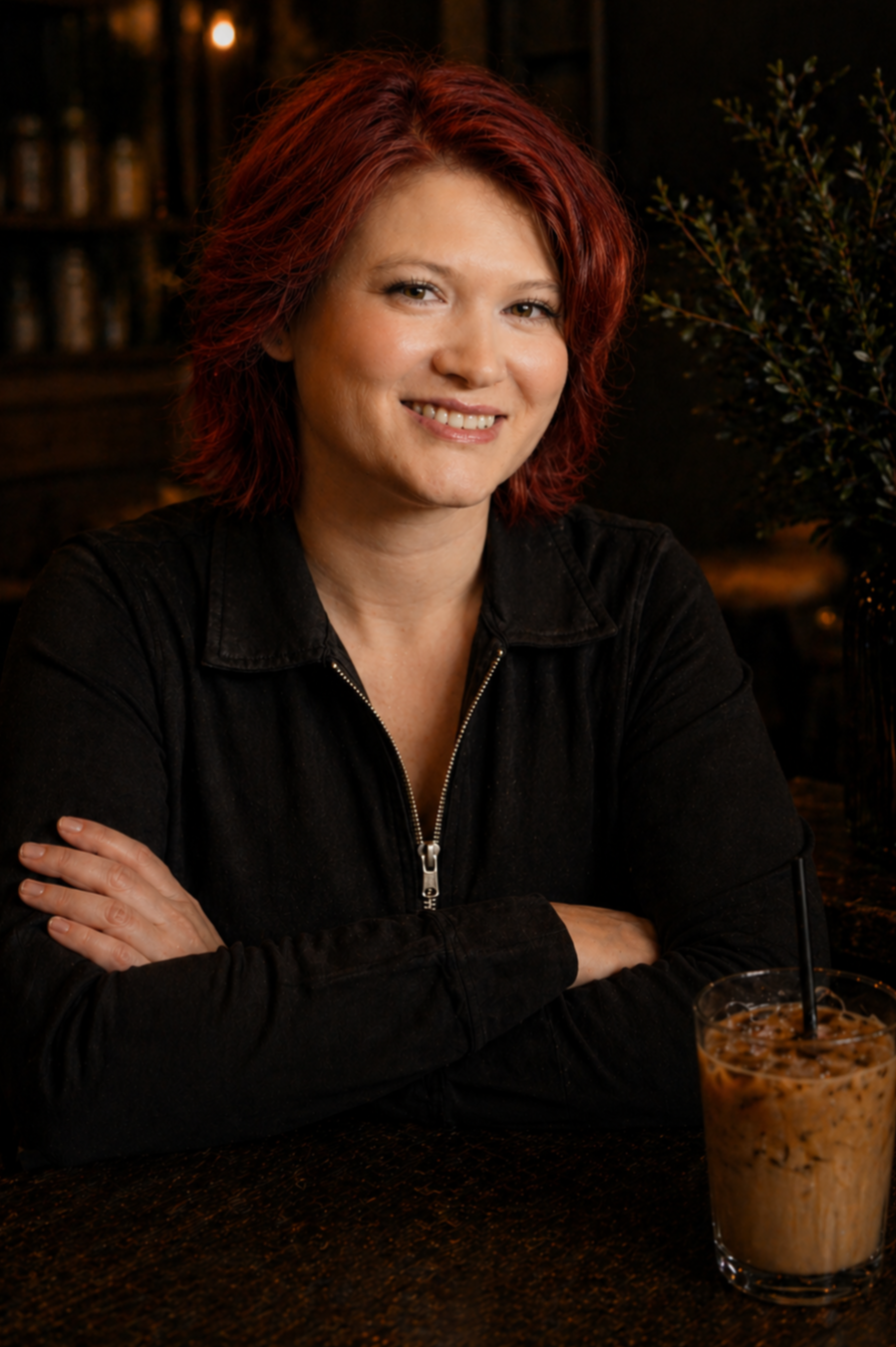 Woman with red hair smiling at a wooden table with an iced coffee drink in a glass.