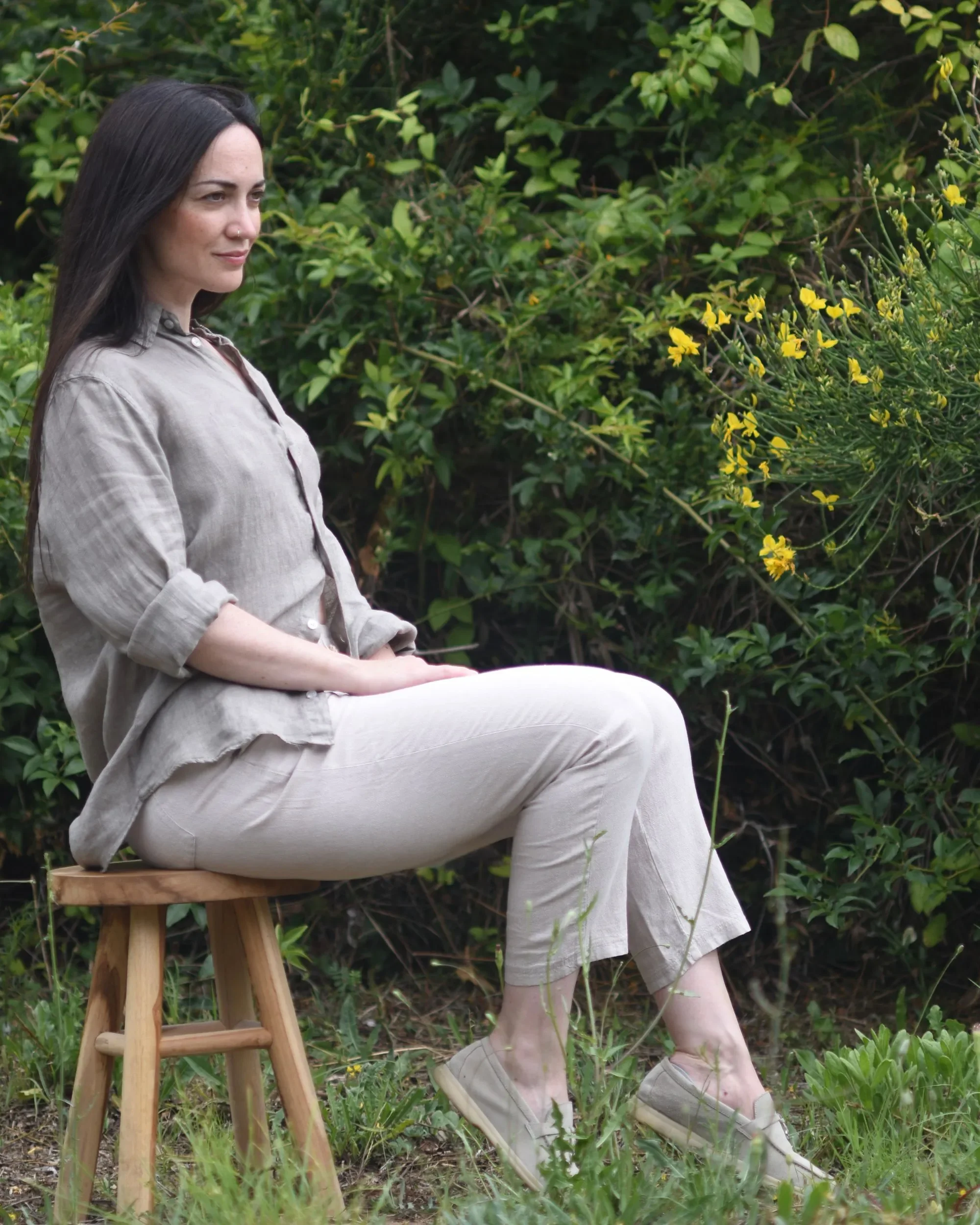 Portrait of artist Francesca V Kranz sitting on a wooden stool in a natural garden setting, representing stillness and contemporary aesthetics.