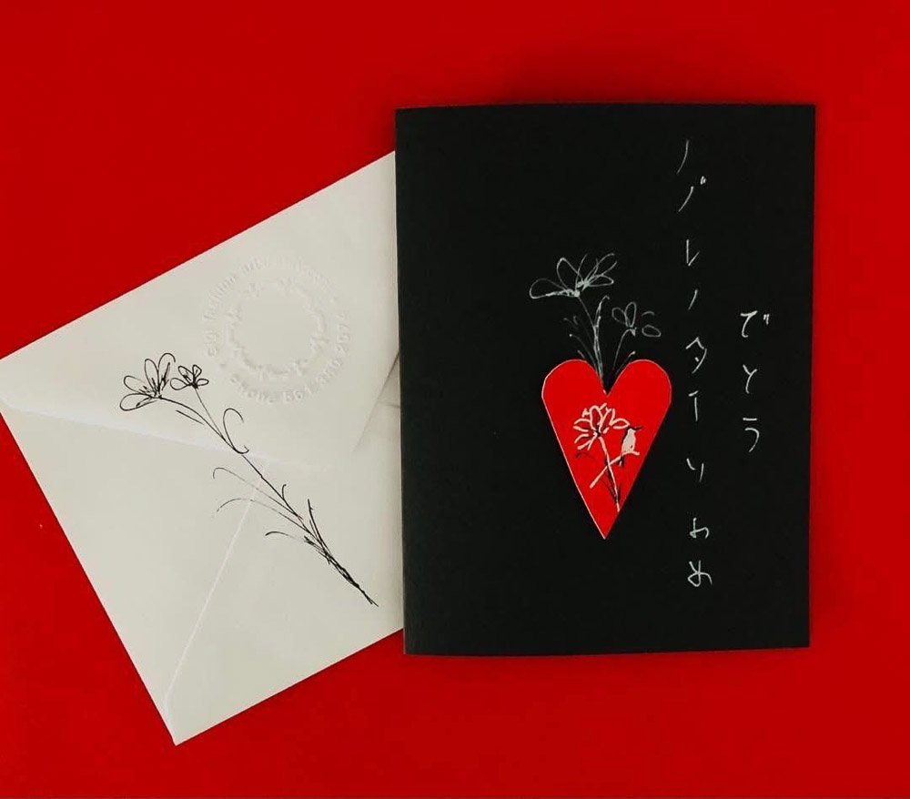 A white envelope with a black plant drawing, placed next to a black greeting card with a red heart, white flower and plant illustration, and Japanese text on a red background.