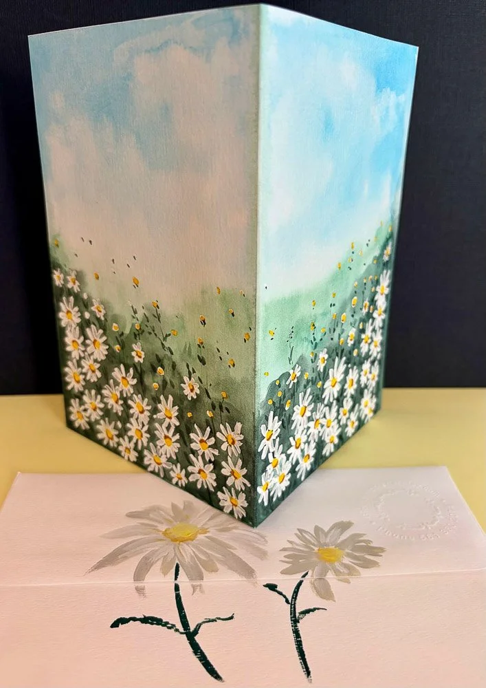 A folding card with a painted landscape of daisies and sky, positioned on a flat surface with a pink floral-patterned envelope in front.