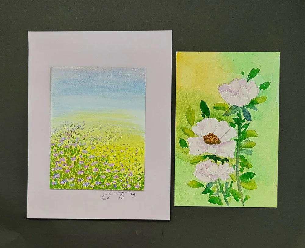 Two watercolor paintings of spring flowers and fields. One depicts a lavender and yellow flower field under a blue sky, the other shows pink flowers with green leaves on a green background.