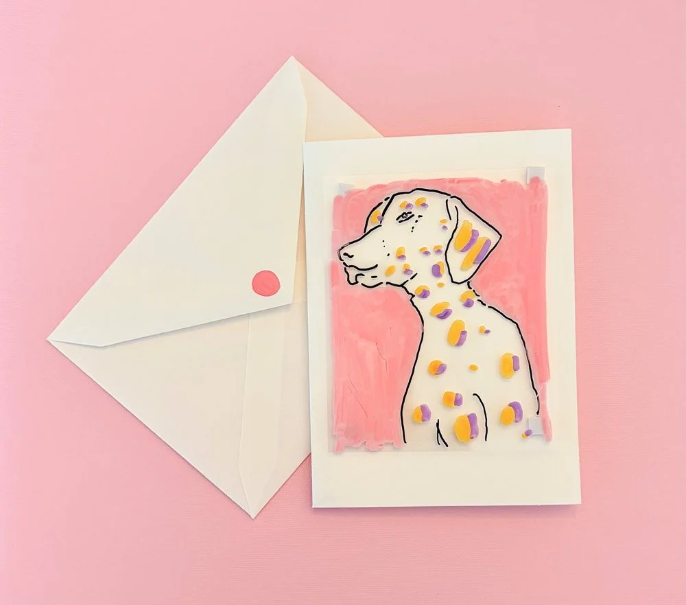 A greeting card featuring a line art illustration of a dog with colorful spots, painted on a pink background, placed next to a white envelope with a pink wax seal on a pink surface.