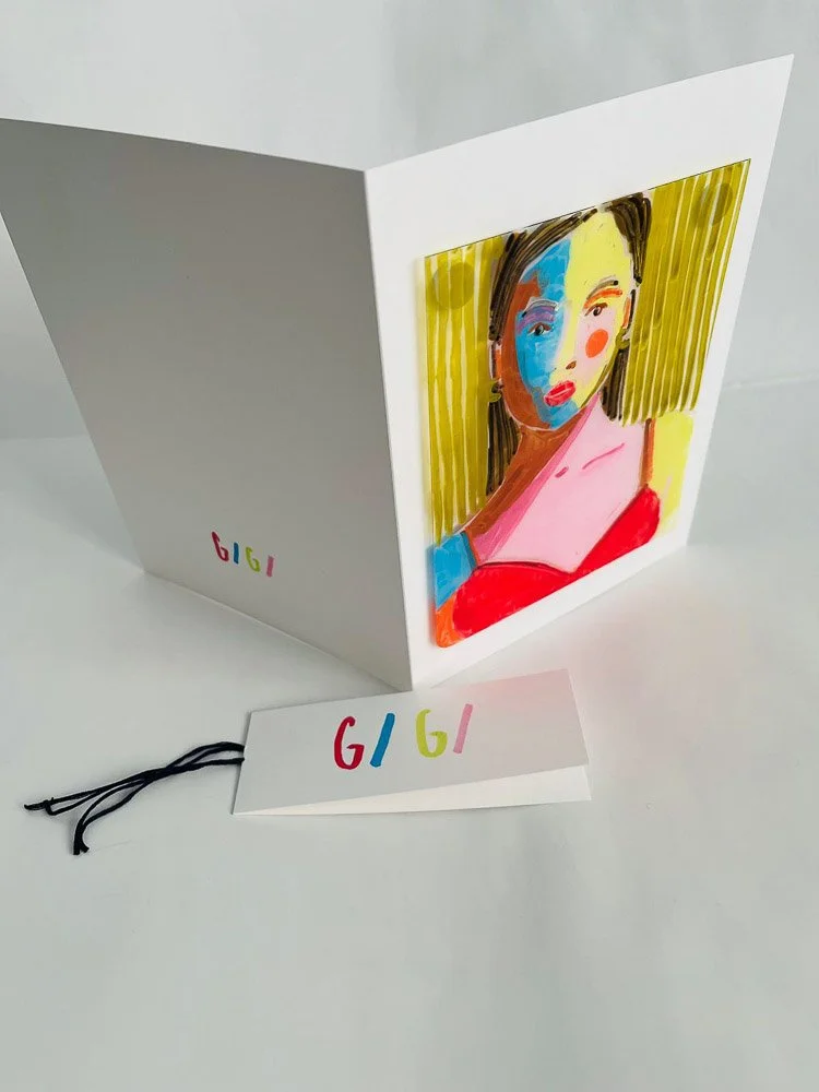 A colorful pop art style portrait of a woman on a white card, with a yellow striped background, inside a white box. The word 'GIGI' is written in multicolored letters on a tag and on the side of the box.