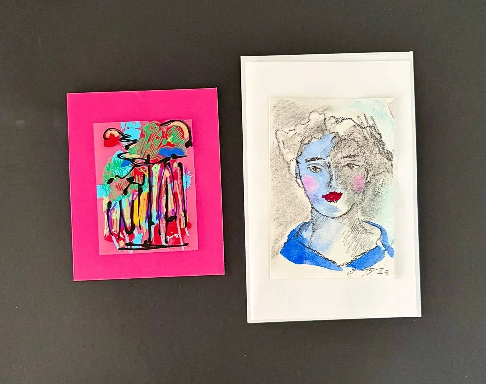 Two framed artworks on a dark wall; the left is an abstract colorful piece with pink background, and the right is a portrait sketch of a woman with blue eyes, red lips, and short hair, in a white frame.