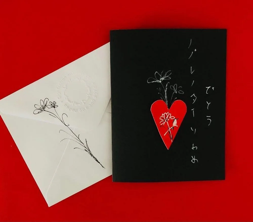 A white envelope with a delicate floral illustration and embossed text, next to a black card featuring a red heart with a floral design and Japanese text, all on a red background.