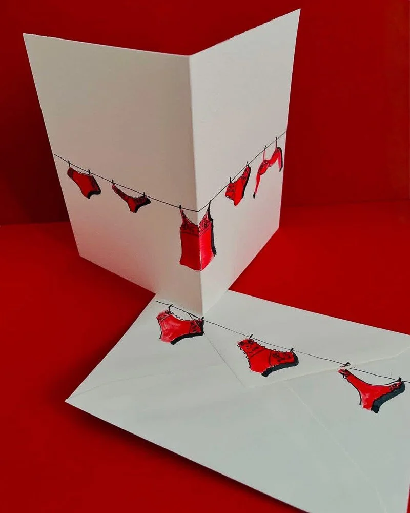 A greeting card with red and black lingerie illustrations hanging on a clothesline on the front, with the matching lingerie inside the card.