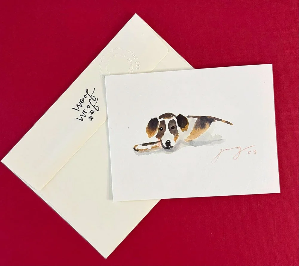 A watercolor painting of a dog lying down, with a white background, placed on a red surface. Next to the painting is a cream-colored envelope with the words 'Woof Woof' and paw prints written on it.