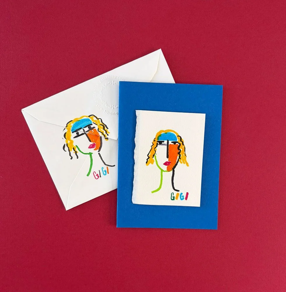 Colorful drawing of a woman's face with blonde hair, orange lips, and black sunglasses, on a white textured paper, placed on a blue card and a white envelope against a red background.