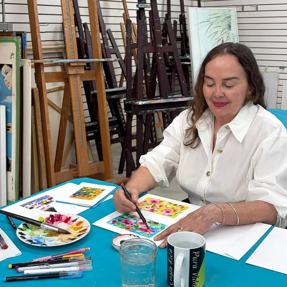 A woman sitting at a table in an art studio, painting colorful abstract art on paper with brushes, with easels and art supplies in the background.