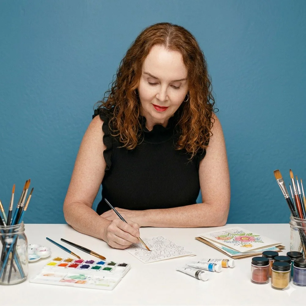A woman with curly red hair sitting at a white table, painting watercolor pictures with brushes and paints in front of her, against a blue wall.