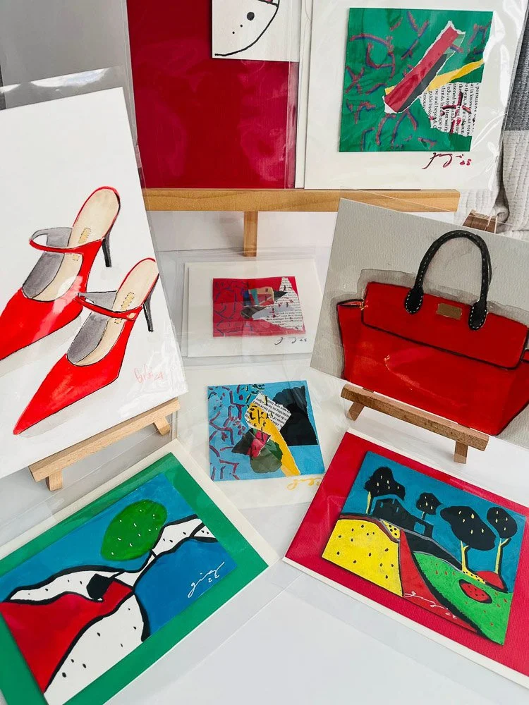 Collection of colorful abstract paintings, some featuring fashion items like red high-heeled shoes and a red handbag, displayed on small wooden easels and a white surface.