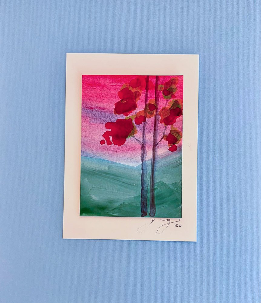 Watercolor painting of a tree with red leaves, against a pink, purple, and green background, framed with a white border.