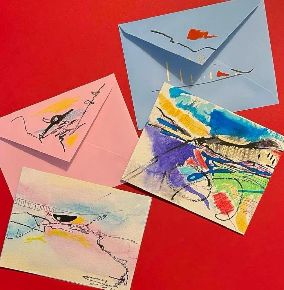 Four abstract greeting cards with colorful, expressive strokes and lines, one with a blue envelope, on a red surface.