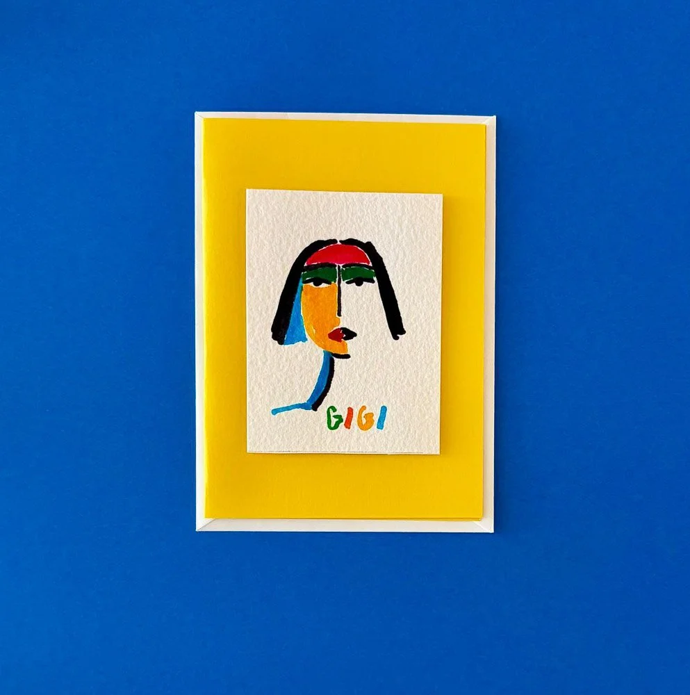 Colorful abstract portrait of a woman with black hair, red lips, and green eyebrows, on textured paper, with the name 'Gigi' written below in various colors, displayed on a yellow-background framed on a blue wall.