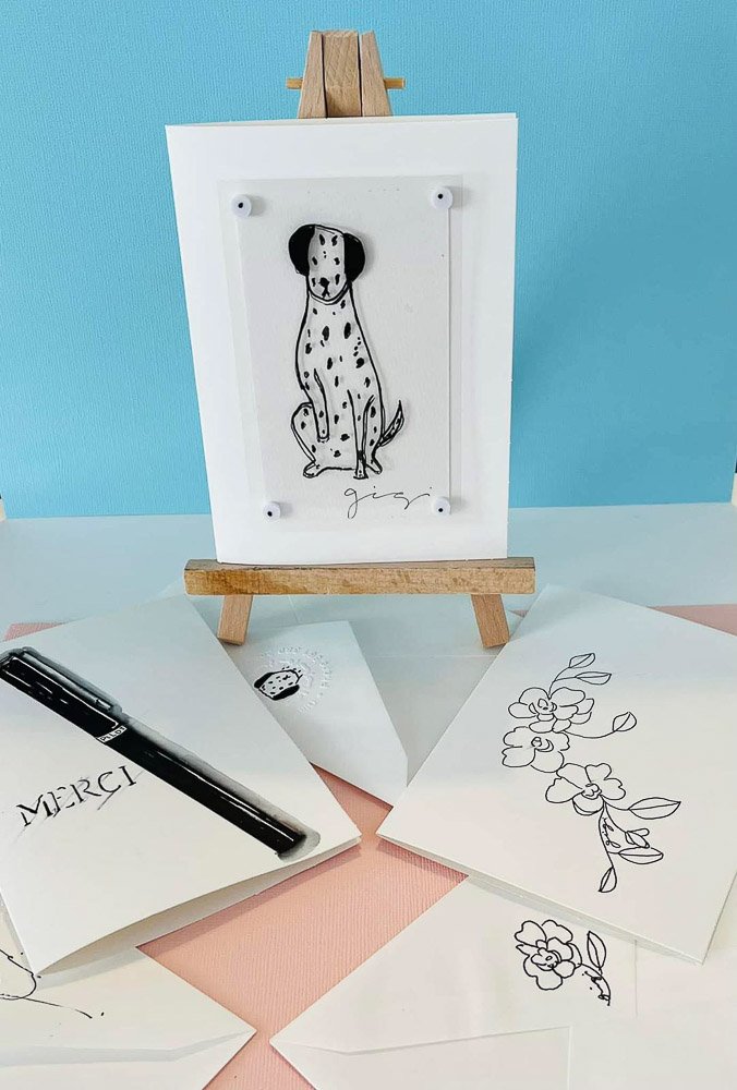 A small wooden easel holding a white framed illustration of a spotted dog with black ears and signature at the bottom. Surrounding the easel are sheets of paper with black floral line drawings, a black marker with a 