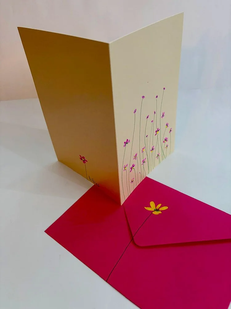 A greeting card with a floral design and a matching pink envelope, featuring simple, colorful flower illustrations, standing upright on a white surface.