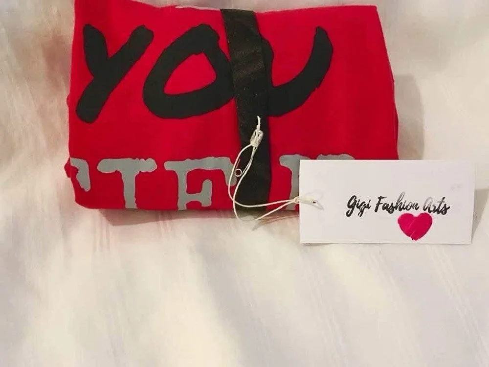 A red gift bag with the words "You" and "The" partially visible in large black and grey letters, tied with a black ribbon, with a white gift tag that says "Gift Fashion Arts" and a red heart.