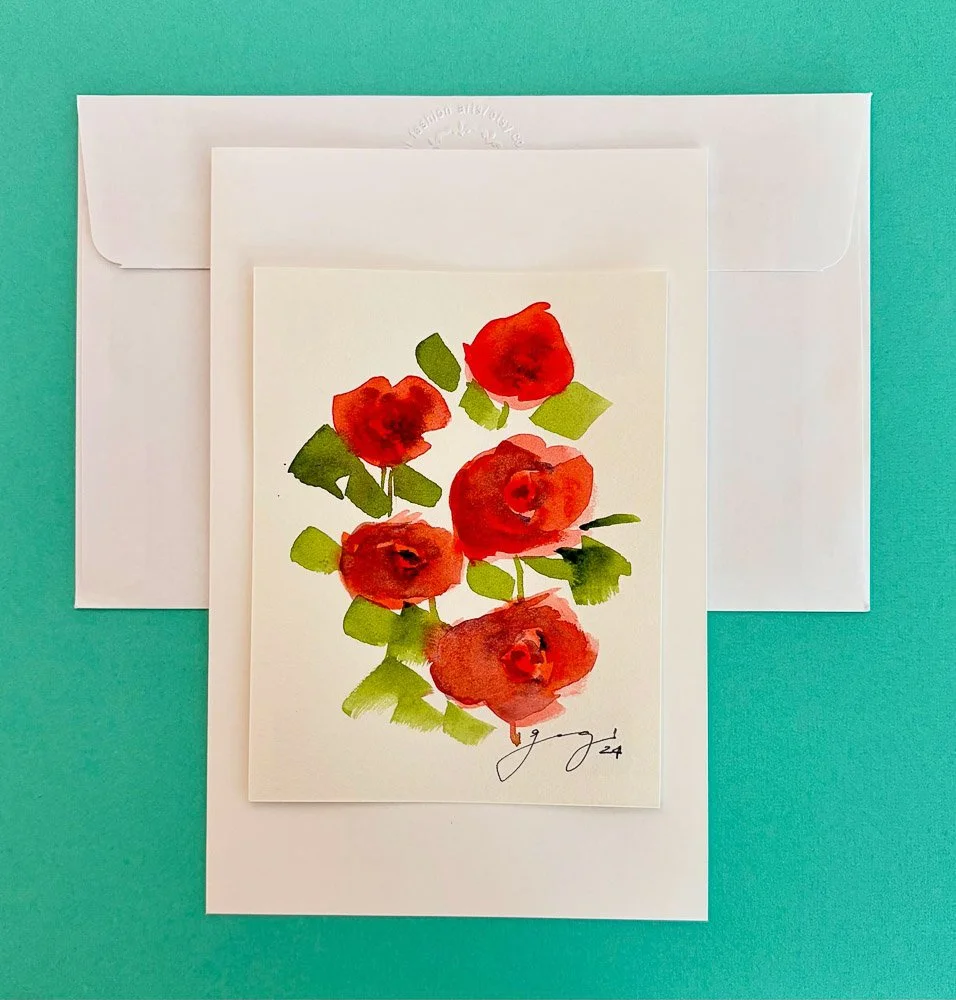 Watercolor painting of red flowers with green leaves on white paper, placed on a white envelope and backing card, set against a teal background.