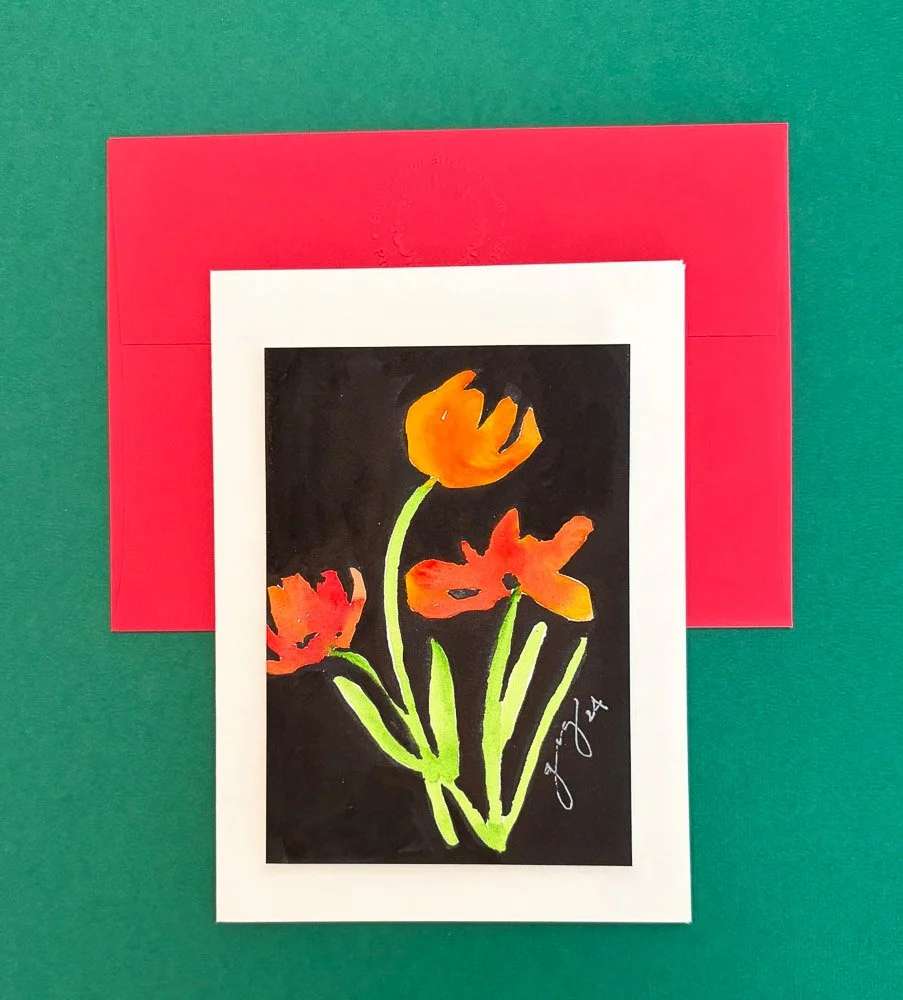 Watercolor painting of orange and red flowers on a black background, signed by the artist, placed on a white mat, with a red envelope and green textured background.