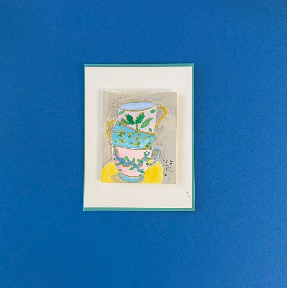 Colorful artwork of stacked teacups with floral designs on paper, mounted on a blue wall.