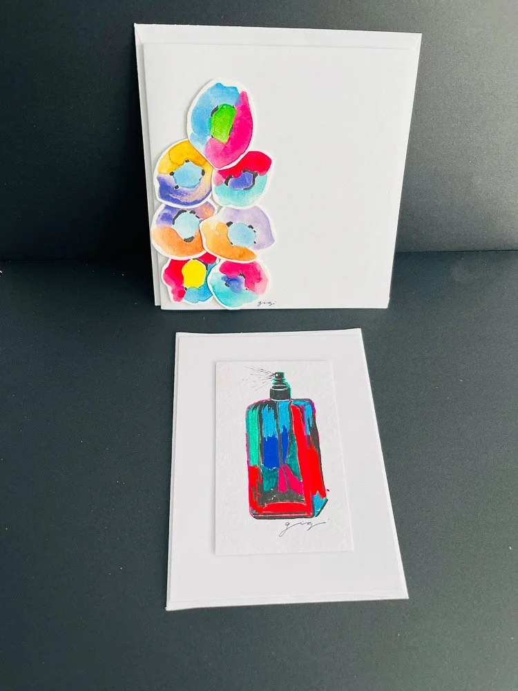 Two pieces of artwork: one in the background with colorful watercolor flowers on a white canvas, and one in the foreground with a colorful illustration of a perfume bottle on white paper.